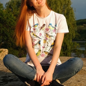 以下が含まれることがあります： カラフルな鳥と花の模様が特徴の白いTシャツ。様々な鳥がピンクのバラの咲く枝に止まっている様子が描かれています。このシャツは半袖で、柔らかい素材で作られています。