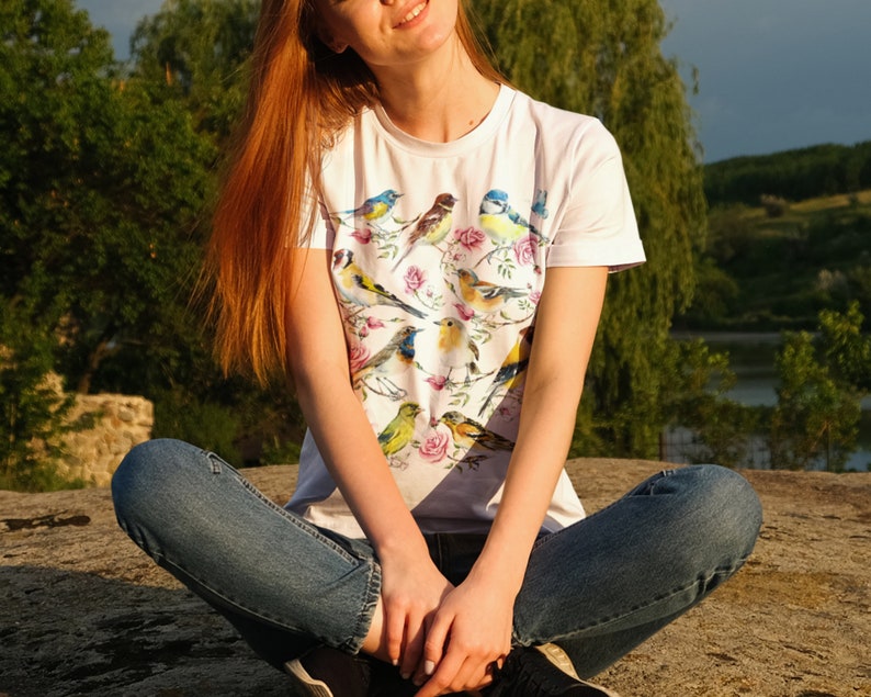 以下が含まれることがあります： カラフルな鳥と花の模様が特徴の白いTシャツ。様々な鳥がピンクのバラの咲く枝に止まっている様子が描かれています。このシャツは半袖で、柔らかい素材で作られています。