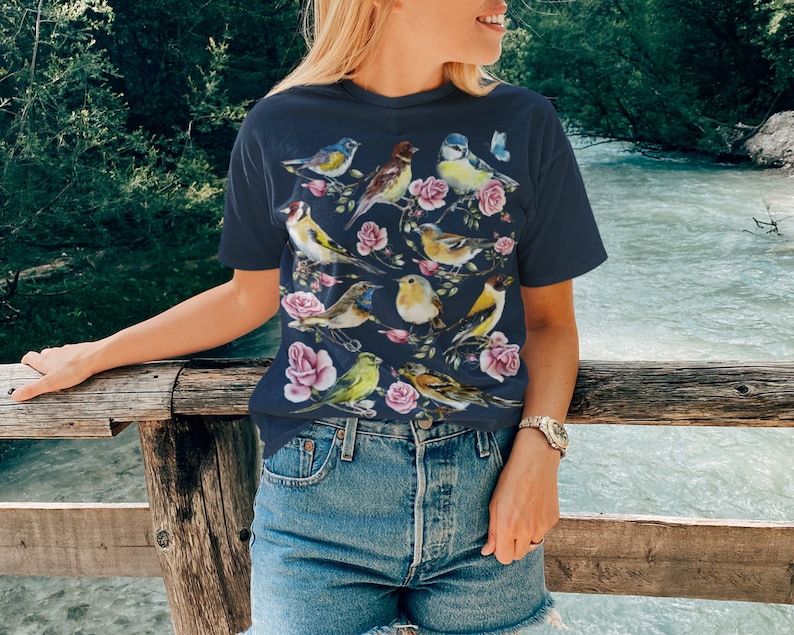 以下が含まれることがあります： ネイビーブルーのTシャツ。水彩画のデザインで、鳥とピンクのバラが描かれています。鳥は黄色、青、茶色、緑の色合いです。カジュアルなスタイルのTシャツです。
