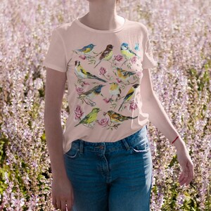 以下が含まれることがあります： カラフルな鳥とバラのデザインが施された、明るいピンク色のTシャツ。半袖のシャツはクルーネックで、ブルージーンズと組み合わせています。鳥は青、黄色、茶色など、さまざまな色で描かれています。
