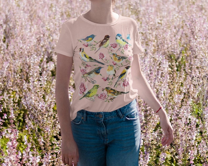 以下が含まれることがあります： カラフルな鳥とバラのデザインが施された、明るいピンク色のTシャツ。半袖のシャツはクルーネックで、ブルージーンズと組み合わせています。鳥は青、黄色、茶色など、さまざまな色で描かれています。
