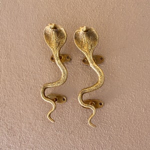 Könnte beinhalten: Zwei goldfarbene Türklinken in Form von Kobras mit aufwendigen Details. Die Griffe sind an einer hellbraunen Wand montiert.