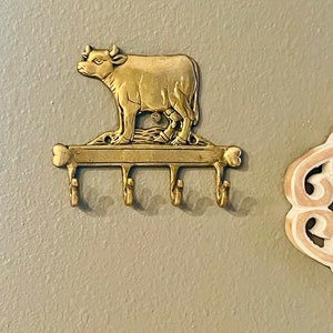 Könnte beinhalten: Ein Schlüsselhalter aus Messing mit einem Kuh-Design. Der Halter hat vier Haken und ist an einer Wand montiert.