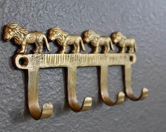 Könnte beinhalten: Goldener Metall-Kleiderhaken mit vier Haken in Form von Löwenköpfen. Der Haken ist an einer grauen Wand montiert.