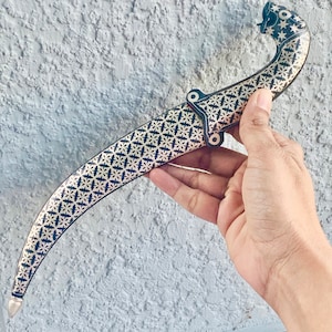May include: A black and silver dagger with a decorative pattern on the blade. The dagger has a curved handle with a stylized animal head.