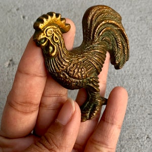May include: A gold-colored metal rooster figurine with intricate details. The rooster is standing on one leg with its wings spread out.