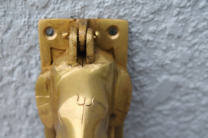 May include: A gold-colored metal door knocker shaped like an elephant's head with a trident symbol on the forehead.