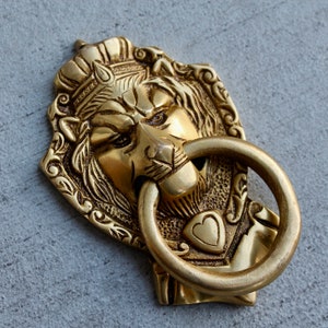 May include: A gold lion's head door knocker with intricate details and a ring handle. The lion is wearing a crown and has a heart-shaped design on its chest.
