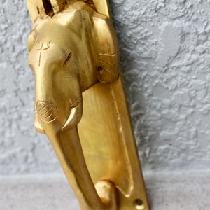 May include: A gold-colored door knocker shaped like an elephant's head with a trident symbol on the forehead. The knocker is mounted on a rectangular plate.