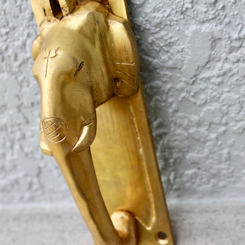 May include: A gold-colored door knocker shaped like an elephant's head with a trident symbol on the forehead. The knocker is mounted on a rectangular plate.