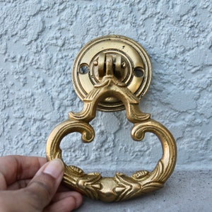 Pu&ograve; includere: Un batacchio per porta dorato con un design decorativo e ornato. Il batacchio ha la forma di un cuore con un'impugnatura ad anello. Il batacchio &egrave; fissato a una piastra rotonda decorativa.