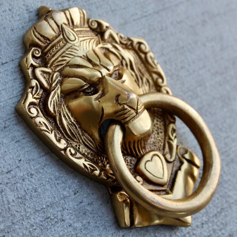 May include: A gold lion's head door knocker with intricate details and a ring pull. The lion's head is surrounded by a decorative frame.
