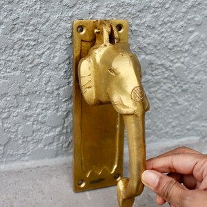May include: A gold-toned door knocker shaped like an elephant's head with a detailed design. The knocker is attached to a rectangular plate with a decorative edge.