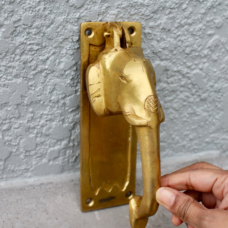 May include: A gold-toned door knocker shaped like an elephant's head with a detailed design. The knocker is attached to a rectangular plate with a decorative edge.
