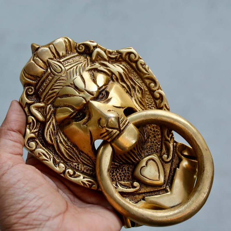 May include: A brass door knocker with a lion's head design. The lion's head is detailed with a crown and intricate carvings. The knocker has a ring for pulling.