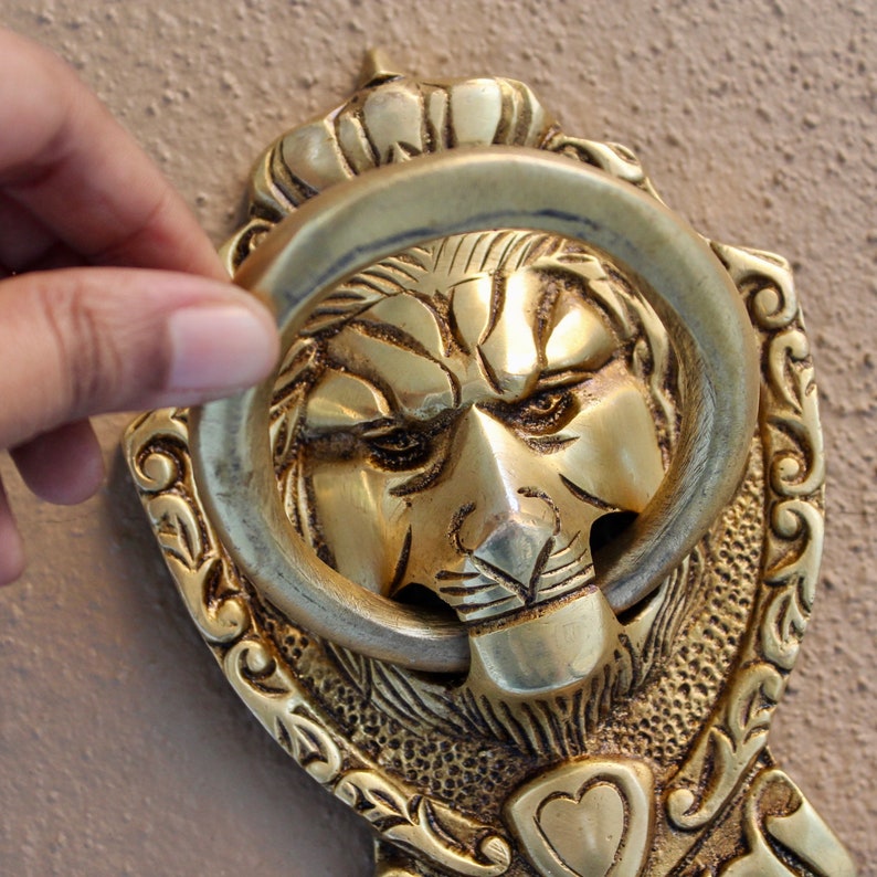 May include: A gold door knocker with a lion's head design. The knocker has a ring handle and intricate details.