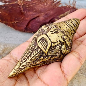 May include: A gold-colored brass conch shell with an intricate design of an elephant head. The shell is decorated with detailed carvings and has a pointed tip.