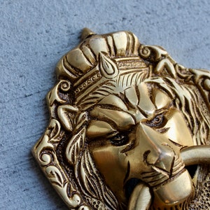 May include: A gold lion head door knocker with intricate details. The lion is wearing a crown and has a ring for knocking.
