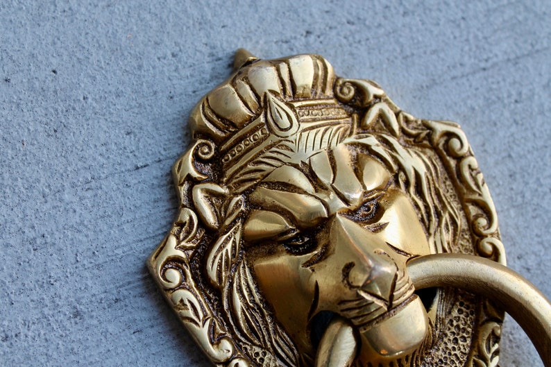May include: A gold lion head door knocker with intricate details. The lion is wearing a crown and has a ring for knocking.