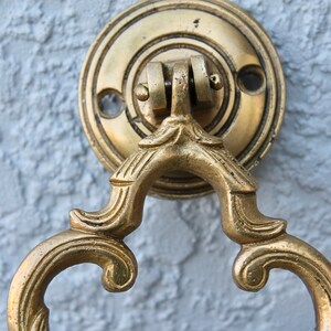 Pu&ograve; includere: Un batacchio per porta dorato con un designo decorativo e ornato. Il batacchio &egrave; attaccato a una piastra rotonda in ottone.