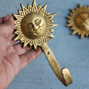 May include: A pair of gold-toned metal sun-shaped hooks with a smiling face design. The hooks are attached to a wall.