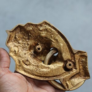 May include: The back of a gold-colored metal door knocker shaped like a hand with a ring. The knocker has two holes for mounting.