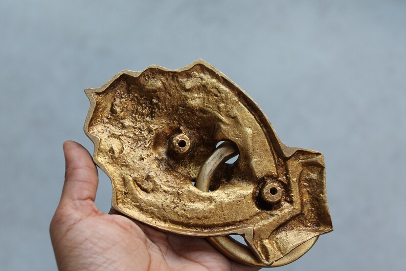 May include: The back of a gold-colored metal door knocker shaped like a hand with a ring. The knocker has two holes for mounting.