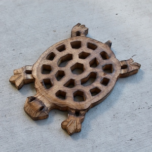 Op de afbeelding: Een houten onderzetter in de vorm van een schildpad met een natuurlijke afwerking. De onderzetter heeft een gedetailleerde gravure van een schildpad met een patroon op de schild en uitgestrekte vinnen. De onderzetter meet ongeveer 25 cm in lengte en breedte.