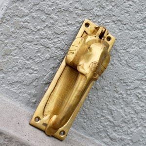 May include: A gold-toned door knocker shaped like an elephant's head with a detailed design. The knocker is mounted on a rectangular plate.