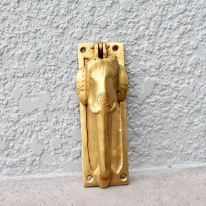 May include: A gold-colored door knocker shaped like an elephant's head. The knocker is mounted on a rectangular plate with two holes for screws.