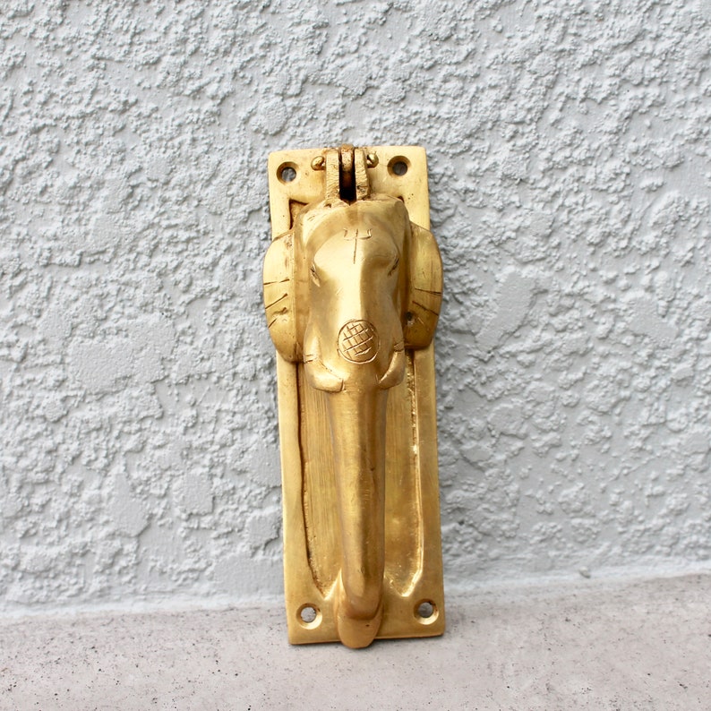 May include: A gold-colored door knocker shaped like an elephant's head. The knocker is mounted on a rectangular plate with two holes for screws.