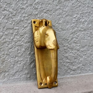 May include: A gold-colored brass door knocker shaped like an elephant's head. The knocker is mounted on a rectangular plate.