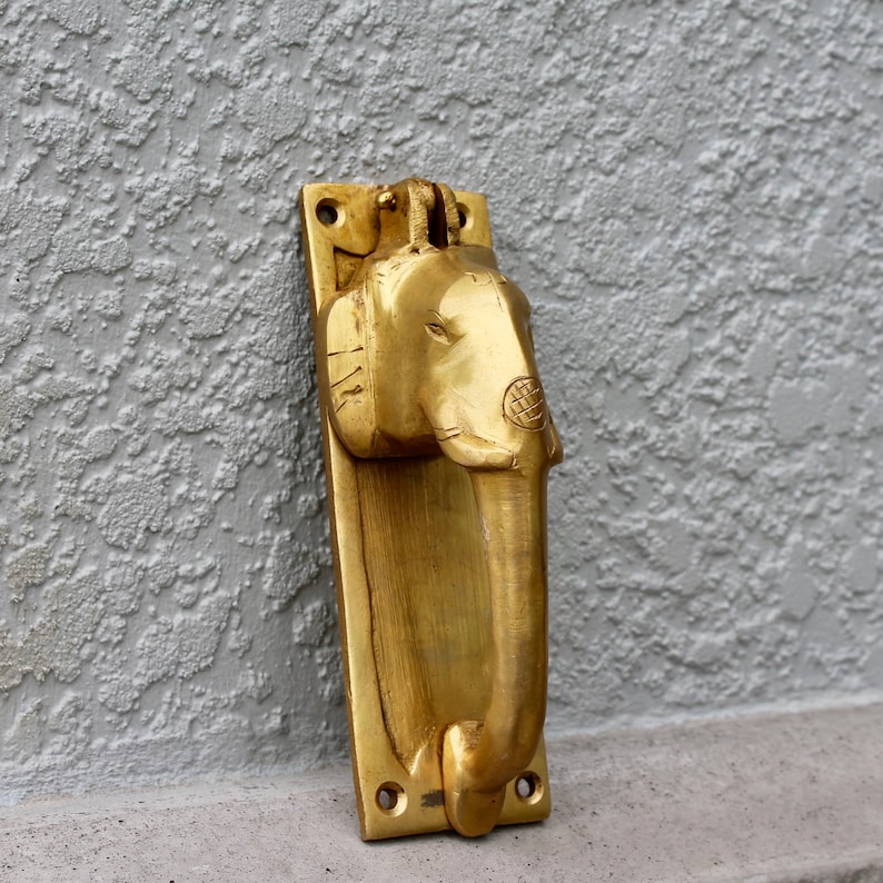 May include: A gold-colored brass door knocker shaped like an elephant's head. The knocker is mounted on a rectangular plate.