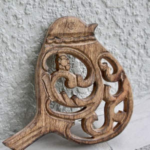 Puede incluir: Un objeto decorativo de madera con forma de pájaro con intrincados detalles tallados. El pájaro tiene una cabeza y cola detalladas, con patrones calados en el cuerpo. La madera tiene un tono marrón cálido natural.