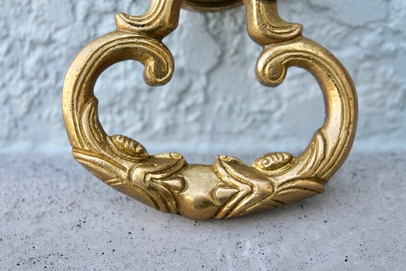 Pu&ograve; includere: Un batacchio per porta dorato ornato con un designo decorativo. Il batacchio ha la forma di un cuore stilizzato con un'impugnatura ad anello.