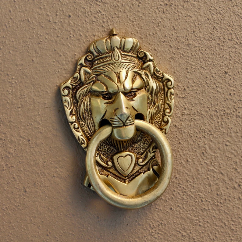 May include: A gold lion head door knocker with a crown and a ring pull. The knocker is detailed with intricate carvings and a heart shape on the chest.