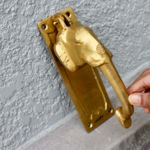 May include: A brass door knocker shaped like an elephant's head with a long trunk. The knocker is mounted on a rectangular brass plate.