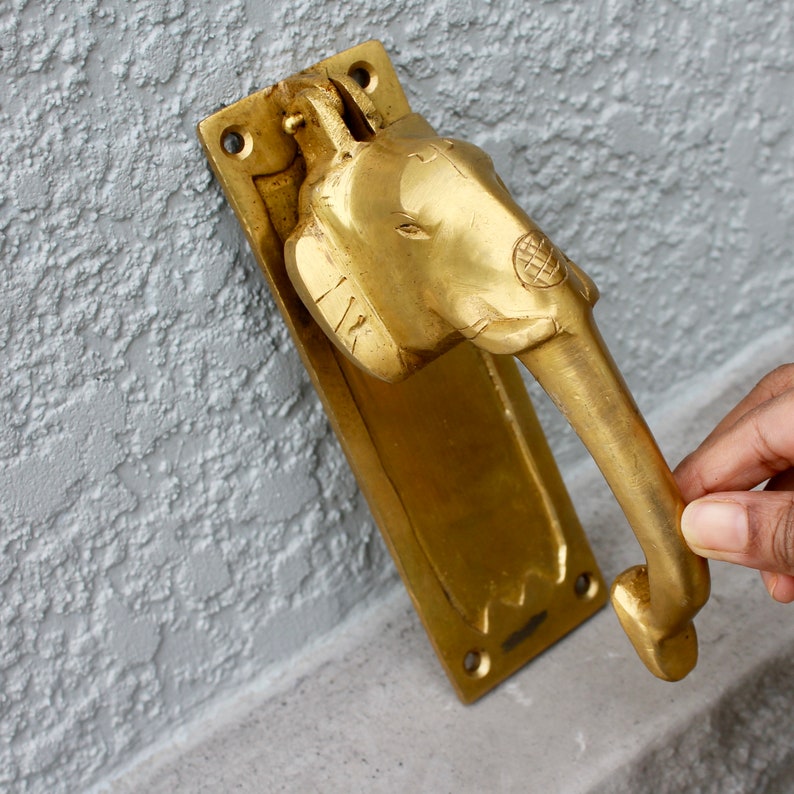 May include: A brass door knocker shaped like an elephant's head with a long trunk. The knocker is mounted on a rectangular brass plate.
