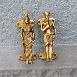 Może przedstawiać: Para złotych metalowych klamki do drzwi w kształcie egipskich postaci. Klamki są ozdobione misternymi rzeźbami i mają tradycyjny design.