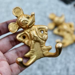 May include: A pair of gold-colored metal wall hooks shaped like a cartoon mouse. The hooks are in a playful pose, with one arm raised and the other extended to form the hook. The mouse has large ears and a smiling face.