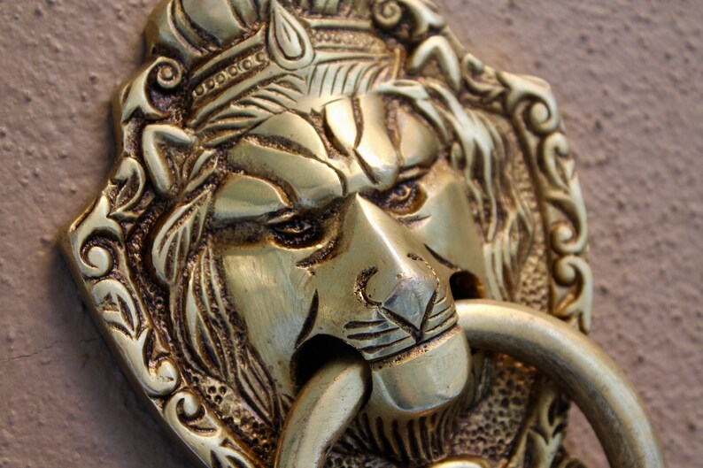 May include: A gold lion's head door knocker with intricate details and a ring for knocking. The lion's head is detailed with a crown and a fierce expression.