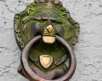 May include: A green and gold lion head door knocker with a ring handle. The lion is wearing a crown and has a heart-shaped shield on its chest.