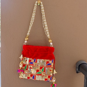 Puede incluir: Una bolsa de cordón de terciopelo rojo con lentejuelas doradas y plateadas y bordados coloridos. La bolsa tiene una correa de cuentas blancas con detalles dorados.