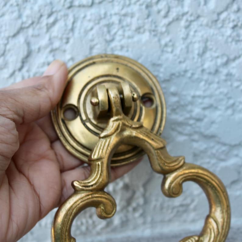 Pu&ograve; includere: Una maniglia per porta dorata con un designo decorativo e ornato. La maniglia ha una base rotonda con un manico ad anello.
