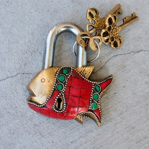 Op de afbeelding: Een messing hangslot in de vorm van een vis met rode en groene mozaïekdetails. Het hangslot heeft een sleutelgat en is bevestigd aan een decoratieve sleutel met een bloemmotief.