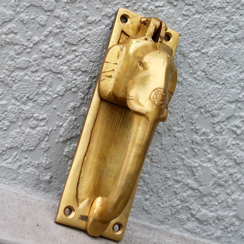 May include: A gold-toned door knocker shaped like an elephant's head. The knocker is mounted on a rectangular plate.