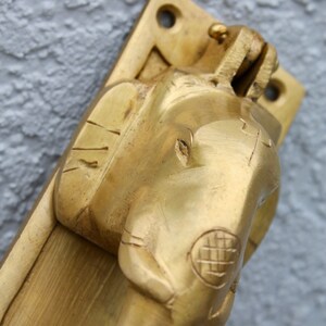 May include: A brass door handle with an elephant design. The handle is attached to a brass plate with a square shape. The elephant's head is facing forward and the trunk is curled up.