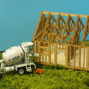 May include: A white toy cement mixer truck with red and blue accents and the word "USA" printed on the side, is parked next to a wooden model house under construction. The house is made of unfinished wood and has a roof frame.
