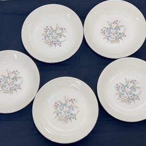May include: A set of five round, off-white ceramic bowls, each featuring a central botanical illustration of light blue berries and tan wheat-like stalks. The bowls have a smooth, glossy finish and a simple, circular shape with a shallow depth.