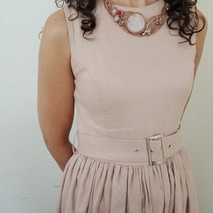May include: A sleeveless light beige dress with a matching belt featuring a silver buckle. The dress is paired with a copper wire-wrapped statement necklace that includes a large central white stone, smaller white beads, and a single red bead.